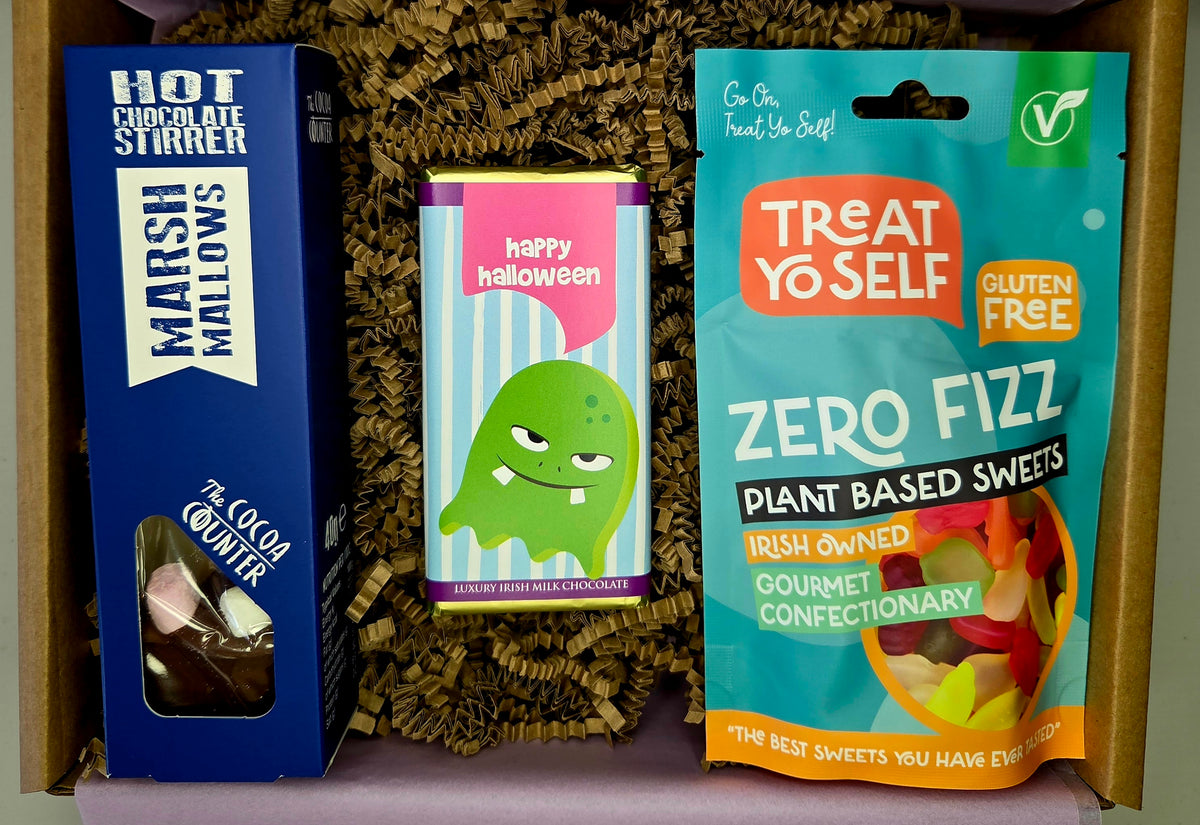 A curated gift box containing three distinct items: a blue box of Hot Chocolate Stirrer Marshmallows, a purple and green striped box of Happy Halloween Fudge Custard, and a teal bag of Treat Yo Self Zero Fizz Plant Based Sweets, all nestled in brown paper shred inside a cardboard box.