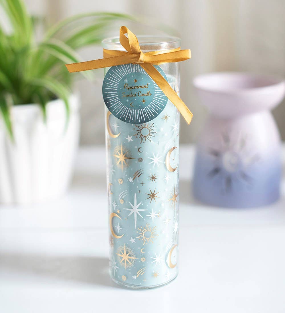 A tall clear glass jar candle with pale blue wax and gold celestial prints, tied with a gold ribbon and a round tag reading 'Happiness is a candle,' set on a white surface with blurred plants and a lavender diffuser in the background.