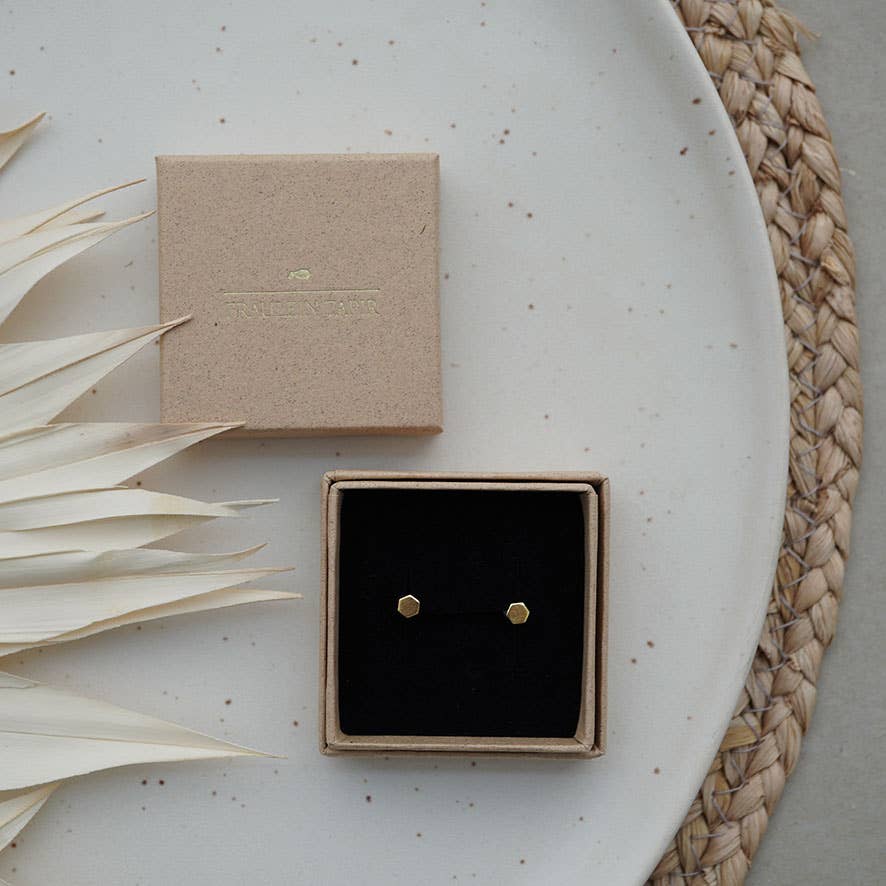 A pair of small gold hexagonal stud earrings displayed in an open square jewelry box with a black insert.