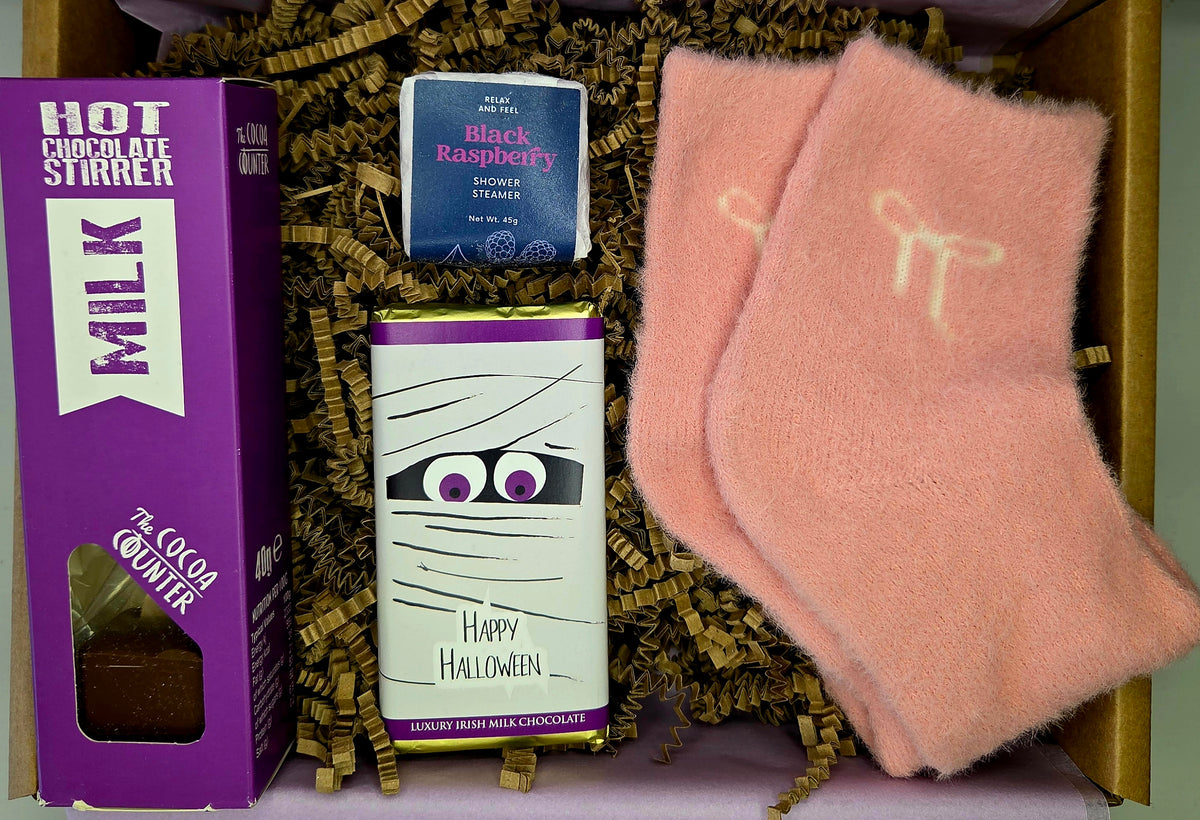 A curated gift box containing a purple box of Hot Chocolate Stirrer milk chocolate, a mummy-themed Happy Halloween chocolate bar, and a pair of soft pink knit socks with a white bow graphic, all nestled in brown paper shred.