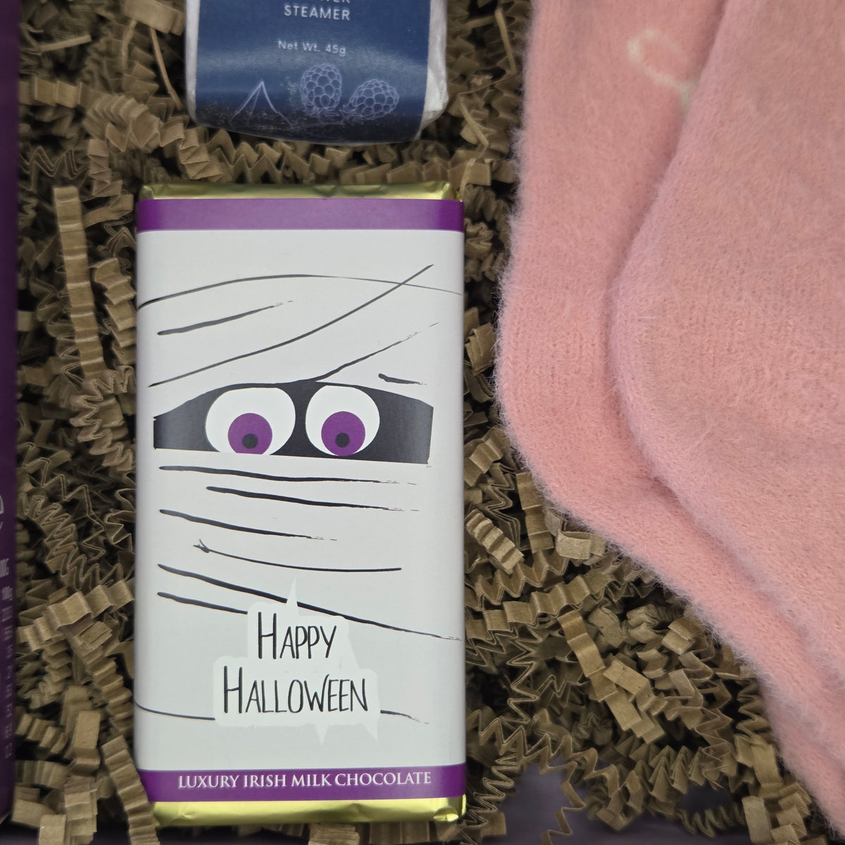 A mummy-themed chocolate bar with purple and gold foil borders and a cartoon face peeking through black wrapping, placed in brown crinkle paper with a pink knit fabric beside it. The bar reads 'Happy Halloween' and 'Luxury Irish Milk Chocolate'.