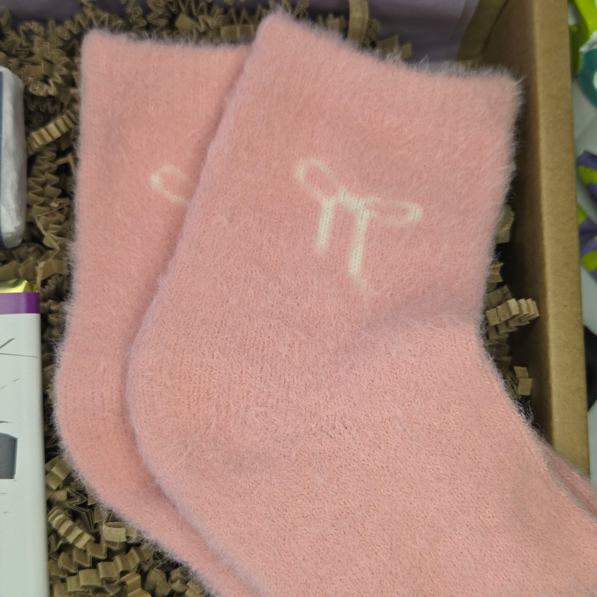 A pair of fuzzy light pink socks with a white bow graphic on the cuff, resting inside a cardboard box filled with brown crinkle paper.