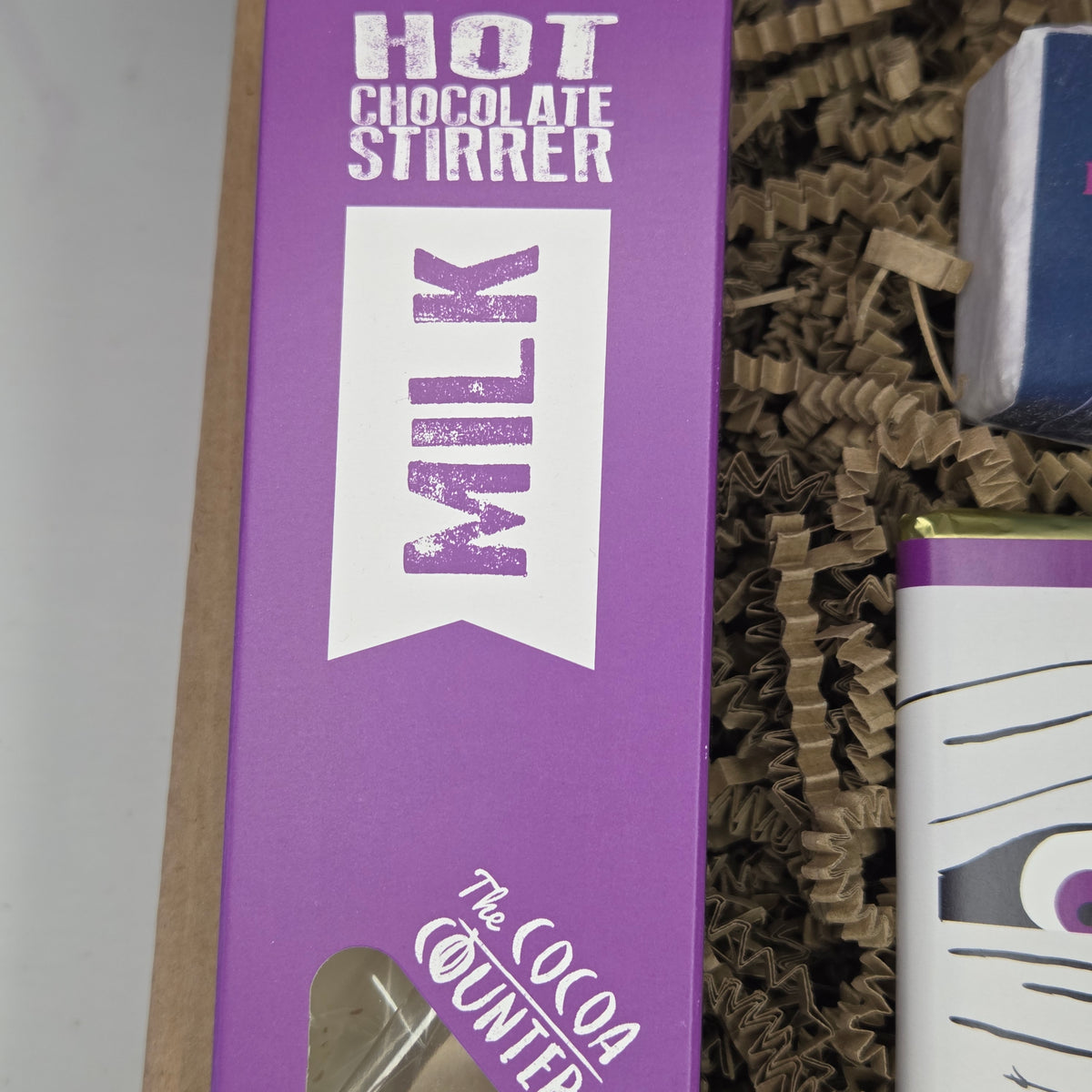 A close-up of a purple box for a hot chocolate stirrer labeled 'MILK' and 'The Cocoa Counter,' placed among brown crinkle paper packaging next to another partially visible product.