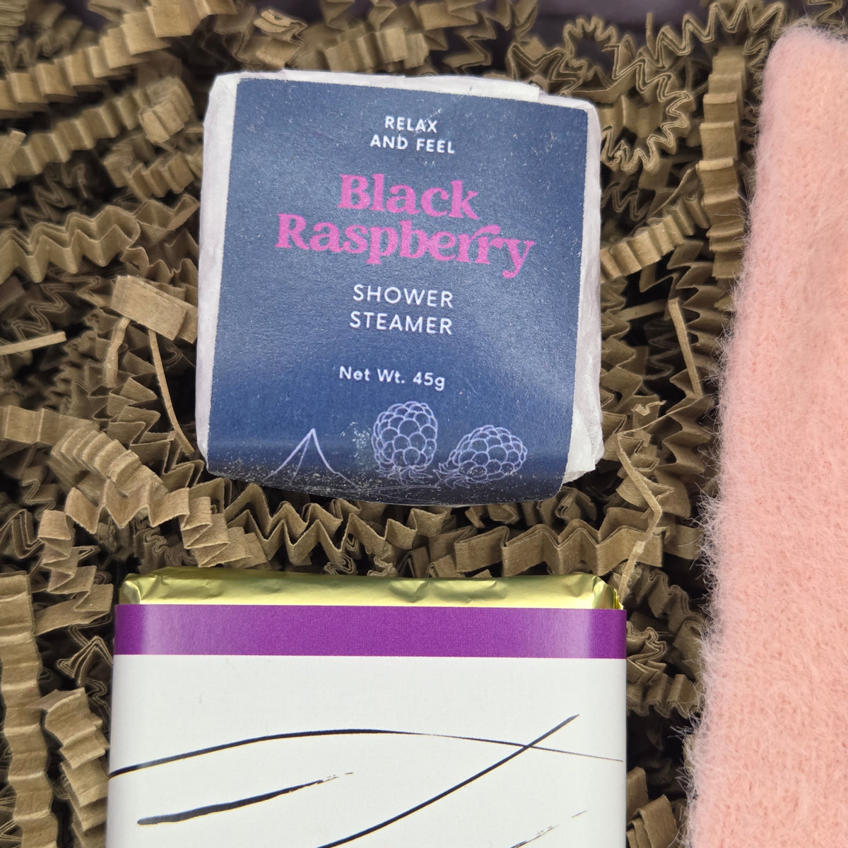 A packaged shower steamer labeled 'Black Raspberry' sits in brown crinkle paper filler, with part of a pink fuzzy item visible at the right edge and another wrapped item below.