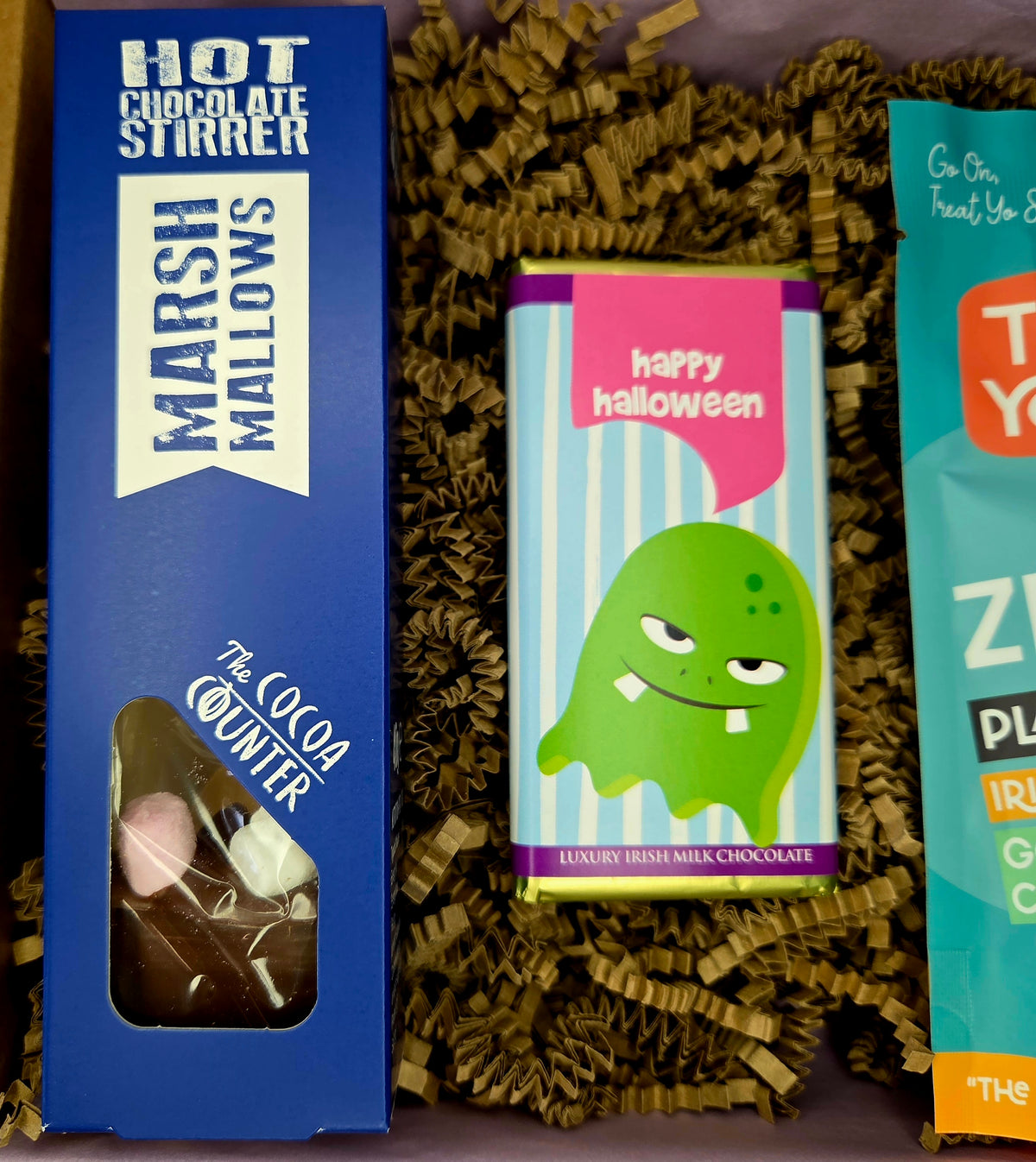 A gift box of chocolates featuring a blue box of 'The Cocoa Counter' Hot Chocolate Stirrer Marshmallows on the left, a green monster-themed 'Happy Halloween' chocolate bar in the center, and a partially visible teal chocolate bar on the right, all nestled in brown crinkle paper.