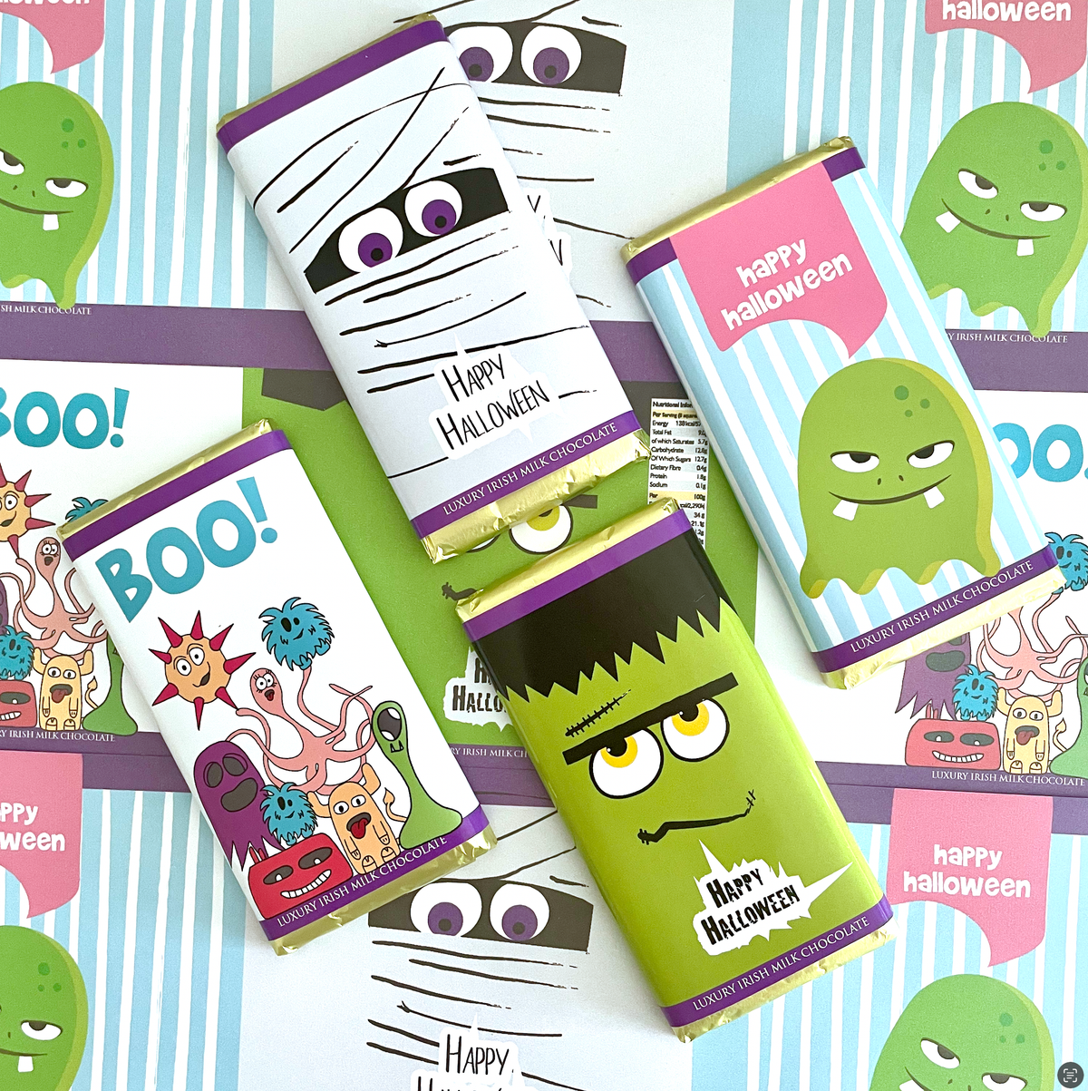 A flat lay of several Halloween-themed chocolate bar wrappers arranged on a striped background. The wrappers feature cartoon monster designs including a mummy, a green monster, a Frankenstein character, and a sun, with text reading 'Happy Halloween' and 'Boo!'.