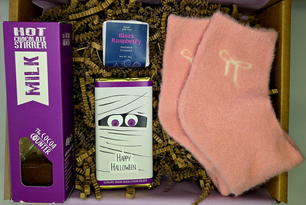 A curated chocolate gift box set containing a purple box of Hot Chocolate Stirrer milk chocolate, a mummy-themed Happy Halloween milk chocolate bar, and a small blue packet of Black Raspberry espresso chocolate, all nestled in brown paper shred next to a pair of fuzzy peach-pink socks.