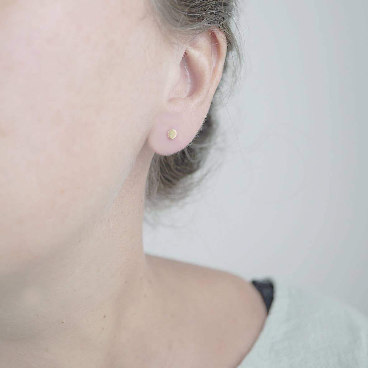 A close-up of a person's ear wearing a tiny round gold stud earring against a soft light gray background.