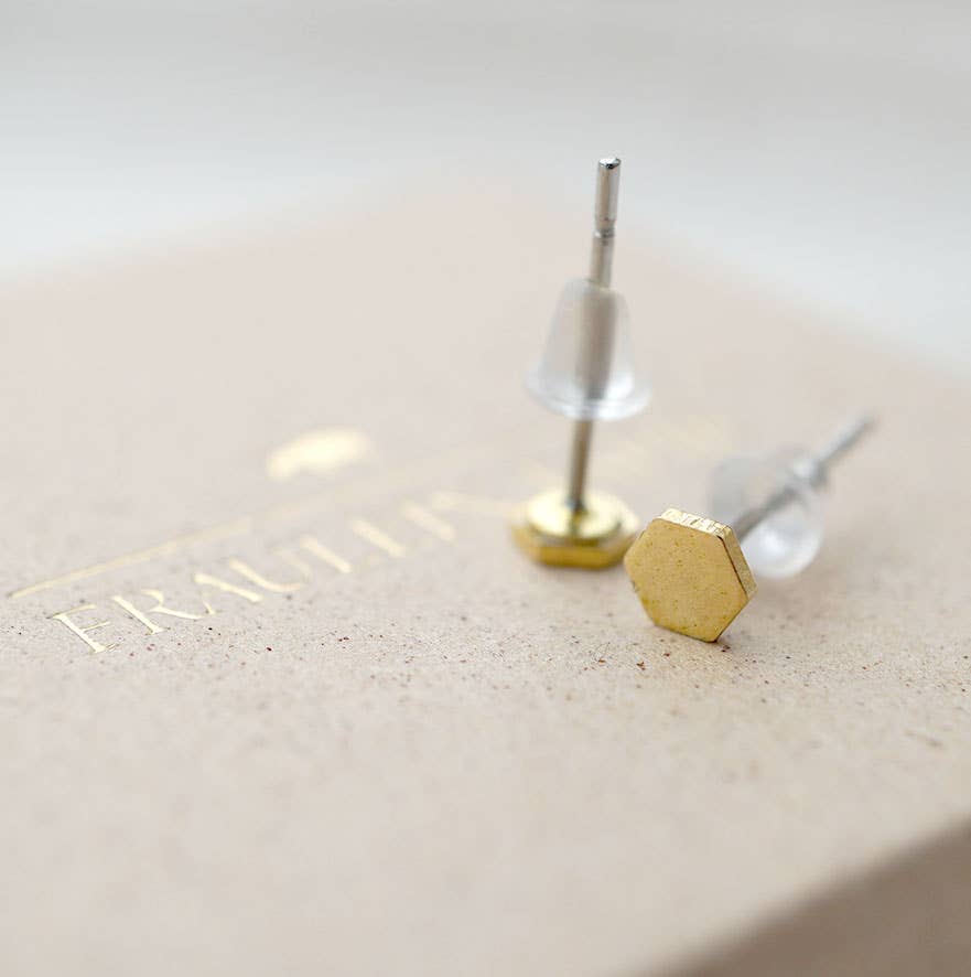 Hexagonal gold stud earrings with clear acrylic backing and silver posts, resting on a textured beige surface with gold foil branding.