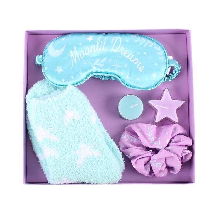 A sleep-themed gift set in a lavender box, featuring a light blue sleep mask with 'Moonlit Dreams' text, a fluffy pale blue towel, a lavender satin scrunchie, and a small star-shaped candle.