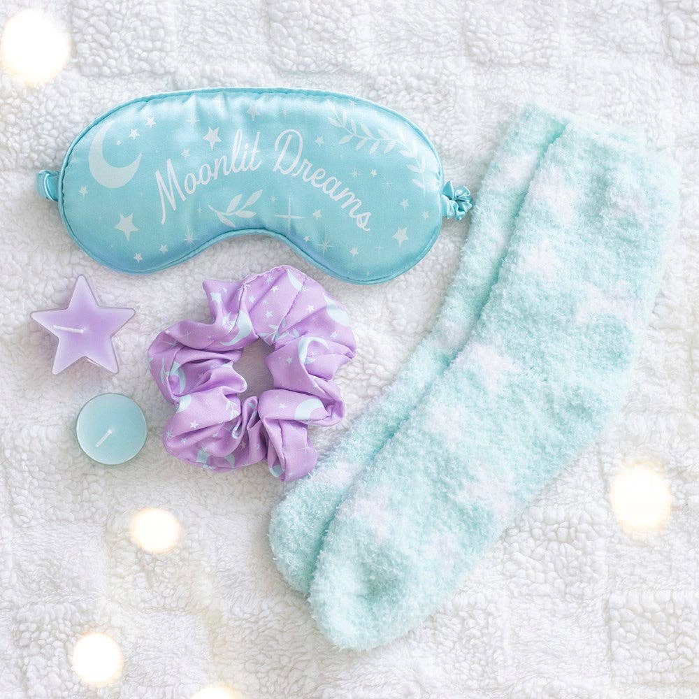 A cozy sleep-themed flat lay featuring a light blue satin sleep mask with 'Moonlit Dreams' text, a fluffy pale blue sock, a lavender satin scrunchie, a lavender star-shaped candle, and a small round candle on a white textured blanket.