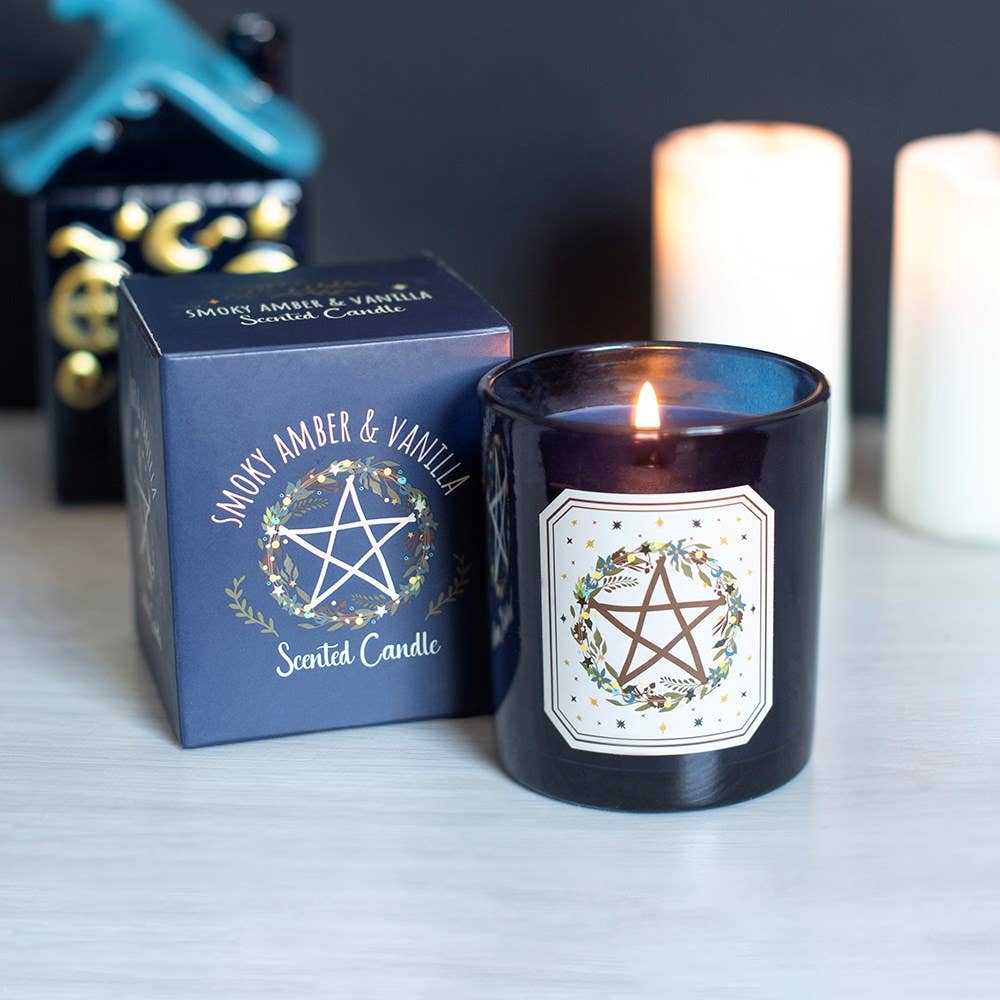 A lit dark blue glass scented candle with a white label featuring a pentagram and floral design, shown next to its matching blue box. The box reads 'Smokey Amber & Vanilla Scented Candle'.