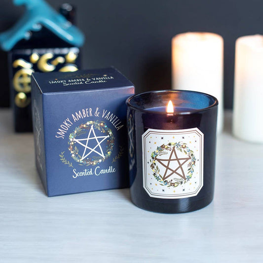 A lit dark blue glass scented candle with a white label featuring a pentagram and floral design, shown next to its matching blue box. The box reads 'Smokey Amber & Vanilla Scented Candle'.