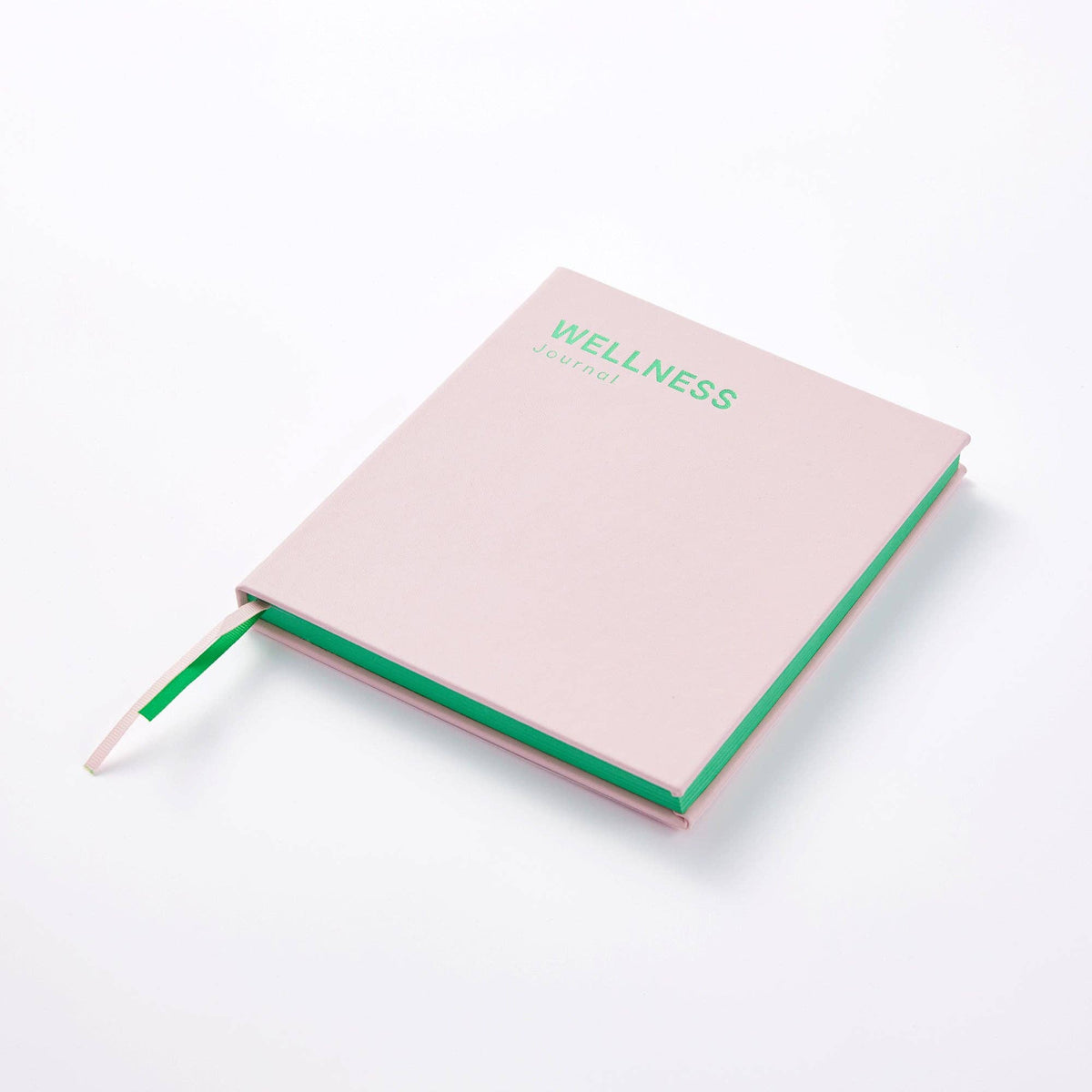 A light pink hardcover journal with a green elastic bookmark ribbon and green text reading 'WELLNESS JOURNAL' on the cover, photographed on a clean white background.