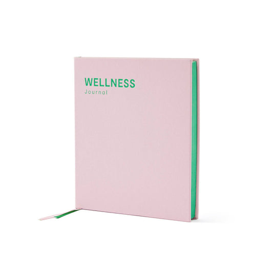 A light pink hardcover journal standing upright on a white background, with the title 'WELLNESS Journal' printed in green on the front cover and a green ribbon bookmark extending from the bottom.